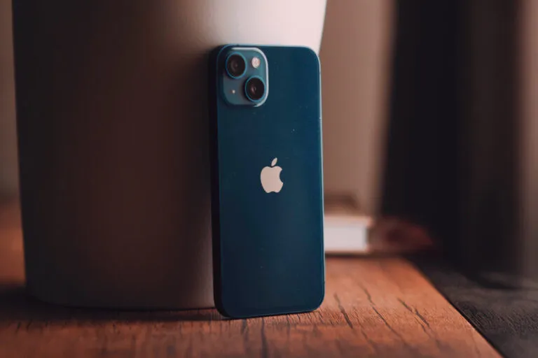 Dos d'un iPhone 13 bleu avec son logo Apple blanc et son double objectif photo sur fond de bois et de tissu
