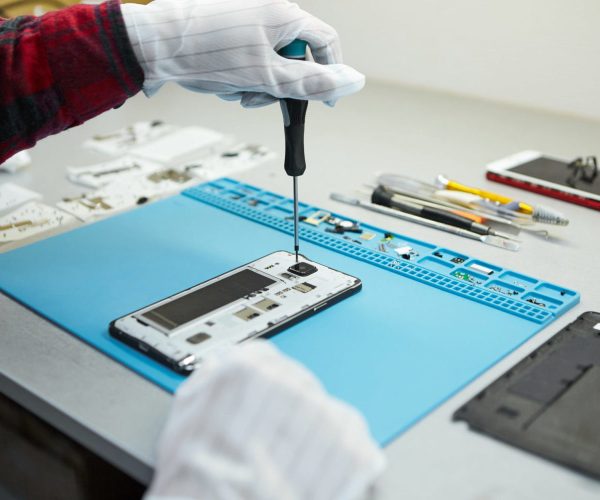 Cropped shot of male technician in antistatic gloves using screwdriver to disassemble borken mobile phone, going to repair motherboard, sitting at his workplace in laboratory with needed equipment